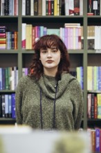 A young female academic pauses thoughtfully in a library, surrounded by colorful bookshelves filled