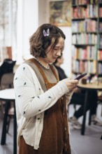 A young woman with a casual style checks her phone in a cozy bookstore. Surrounded by vibrant
