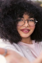 A young latin woman with voluminous curly hair, wearing round glasses and a cozy sweater, smiles