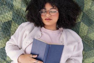 A latin woman with curly hair and glasses peacefully rests on a patterned blanket, holding a book.