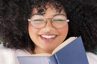 A joyful latin woman with curly hair and large round glasses, holding a book with delight. Her