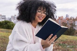 A latin woman with curly hair and glasses enjoys reading a book in an outdoor park setting. She is