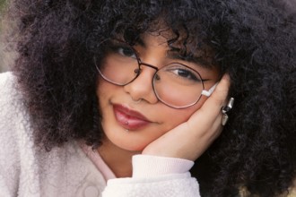 A young woman with curly hair wearing glasses and piercings is captured up-close. Her thoughtful