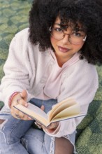 A young latin woman with glasses enjoys reading a book while sitting comfortably. Her casual style