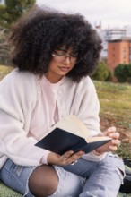 A latin woman with curly hair and glasses enjoys reading a book while seated on grass in a peaceful