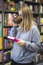 A woman in a gray hoodie drinks coffee and reads a book in a cozy bookstore. The background