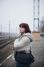 A young woman in a puffer jacket holds a coffee cup, waiting at a train station on a foggy winter