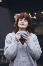A young woman with closed eyes savors a moment with a cup of coffee. She wears a cozy gray sweater,