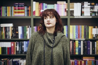 A young woman with curly hair, wearing a green sweater, stands ponderatively near a bookcase filled