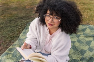 A young latin woman with curly hair and glasses sits on a picnic blanket outdoors, absorbed in