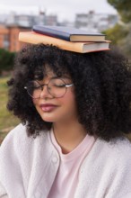 A young latin woman with curly hair and glasses balances two books on her head while outdoors in a