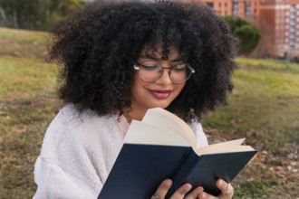 A young latin woman with curly hair and glasses reads a book while sitting on grass in a park. Her
