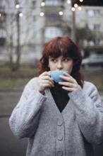 A young woman enjoys a coffee from a blue cup while standing outdoors on cafe terrace. She is