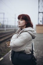 A young woman in a white coat stands at a train station, holding a coffee cup. The scene is foggy,