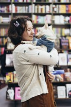 A young woman holds her dog lovingly in a warm bookstore, surrounded by colorful books. The cozy