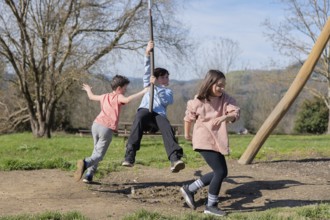 Three friends play joyfully on a zip line in a park, enjoying a sunny day together. Trees and green