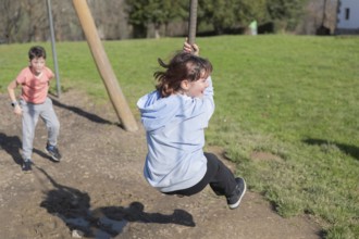 Two friends enjoy playing on a zip line in a sunny park. One child swings while the other waits in