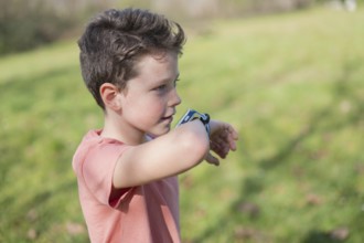 A young boy in a coral shirt examines his smartwatch in a sunny park. The background features