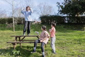 Three friends playfully interact on a wooden picnic table in a sunny park. One child stands