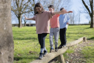 Three friends are balancing on a wooden beam in a park. They are enjoying the sunny day, surrounded
