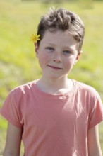 A young child with short hair and a flower tucked behind the ear enjoys a sunny day outdoors. The