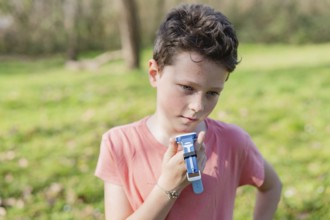 Young boy in pink shirt holding a blue smartwatch in a green park. Sunlit scene with blurred