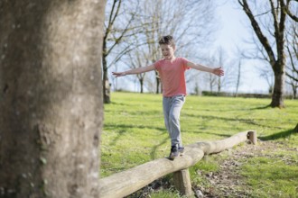 A young boy skillfully balances on a log in a sunlit park. Trees surround him, and there a vibrant