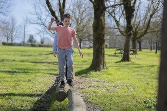 Friends skillfully balances on a log in a sunlit park. The background features lush grass, trees,