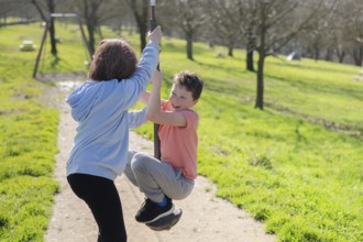 Two friends play joyfully on a zip line in a sunny park. Surrounded by green grass and trees, they