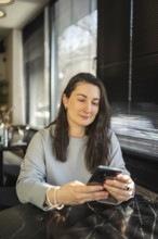 A mid-aged woman focuses intently on her smartphone while sitting at a cafe table bathed in