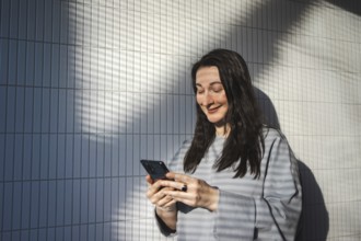 A mid-aged woman enjoys the sunlight as she smiles and engages with her smartphone outdoors against