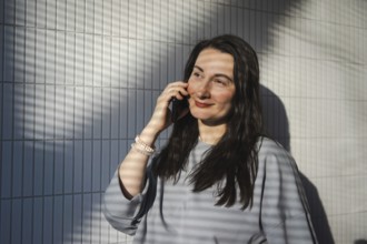 A mid-aged woman enjoys a sunny day at a cafe, chatting on her smartphone against a geometric