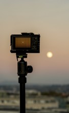 A camera on a tripod silhouetted against a warm sunset sky, overlooking the cityscape of Geneva,