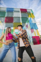 A joyful lesbian couple dances and laughs together in an urban setting. The vibrant glass wall in