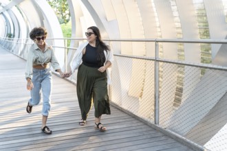 A lesbian couple holds hands as they joyfully walk on a sunlit, modern bridge. Both wearing casual