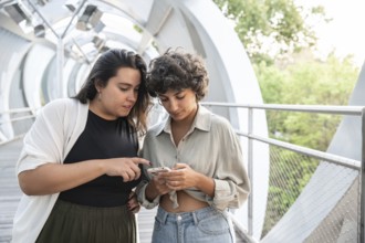 A lesbian couple shares a moment on a contemporary walkway, looking at a smartphone. The scene