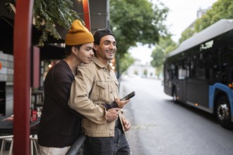 A joyful gay couple embraces by a bus stop. They are smiling, holding a phone, with trees,