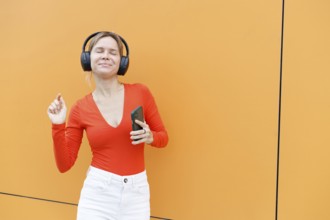 A woman stands against an orange wall in Warsaw, wearing headphones and a red top She holds a