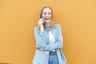 A cheerful woman in a blue shirt talks on her smartphone against an orange background in Warsaw,