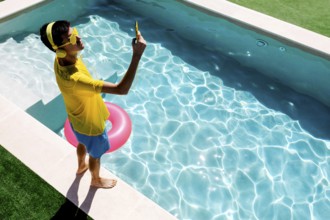 A young teenage boy in vibrant yellow attire takes a selfie by a sparkling blue pool. Sunlight