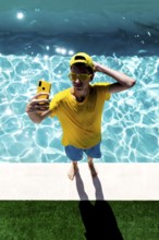 A teenage boy in a yellow shirt, sunglasses, and cap takes a selfie by a bright blue pool. His