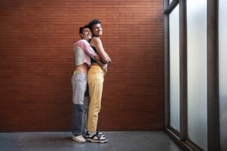 A joyful gay couple embraces lovingly against a brick wall. Their happy expressions and casual