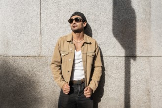 A man in a tan jacket and sunglasses leans against a sunlit concrete wall. The casual attire and