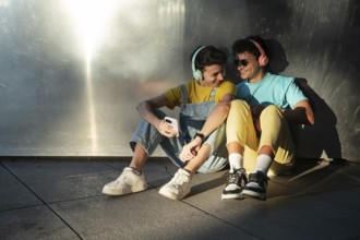A gay couple sits joyfully against a metallic wall, sharing music through headphones. Their