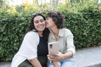 A joyful lesbian couple takes a selfie in a lush park. One partner kisses the other on the cheek,
