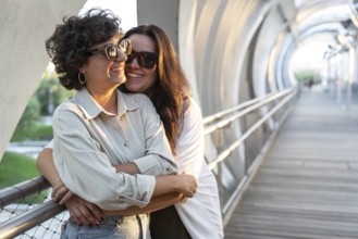 Lesbian couple warmly embracing while standing on a sunlit street bridge. Both wearing sunglasses,