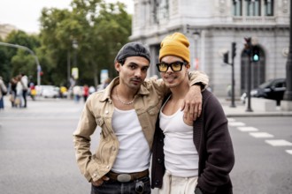 A Colombian gay couple stands joyfully in an urban setting. They wear casual outfits, expressing a
