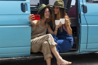 Two lesbians women enjoying a road trip sit by a turquoise van. They smile, capturing the moment