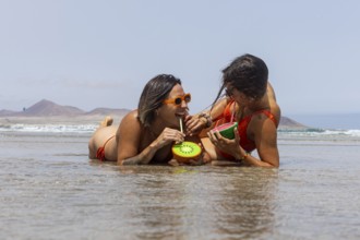 Two lesbians women in swimsuits enjoy refreshing drinks on the beach, laughing and relaxing by the