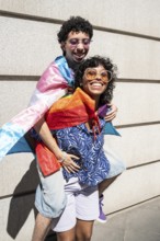 A vibrant, joyful multiethnic LGBTQ+ couple enjoys a sunny day outdoors, wrapped in pride flags.